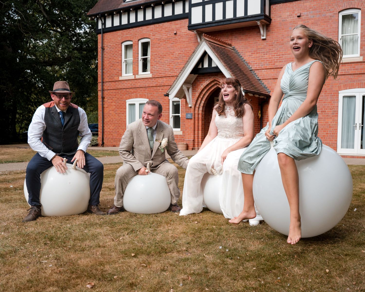 Wedding party sitting on white spherical outdoor seats in front of a red brick Tudor-style manor house.