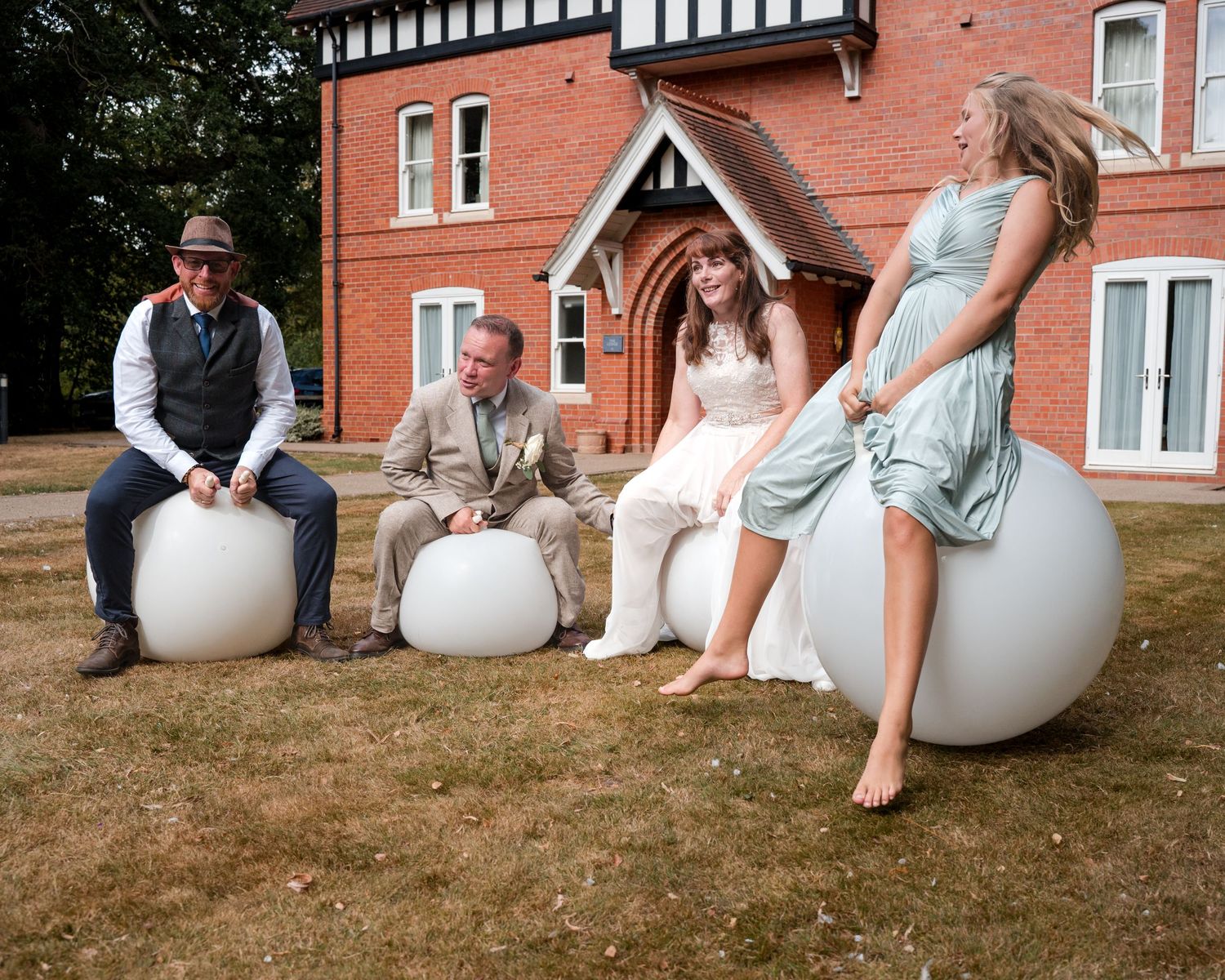 Wedding party sitting on white spherical seats outside a red brick manor house on a sunny day.