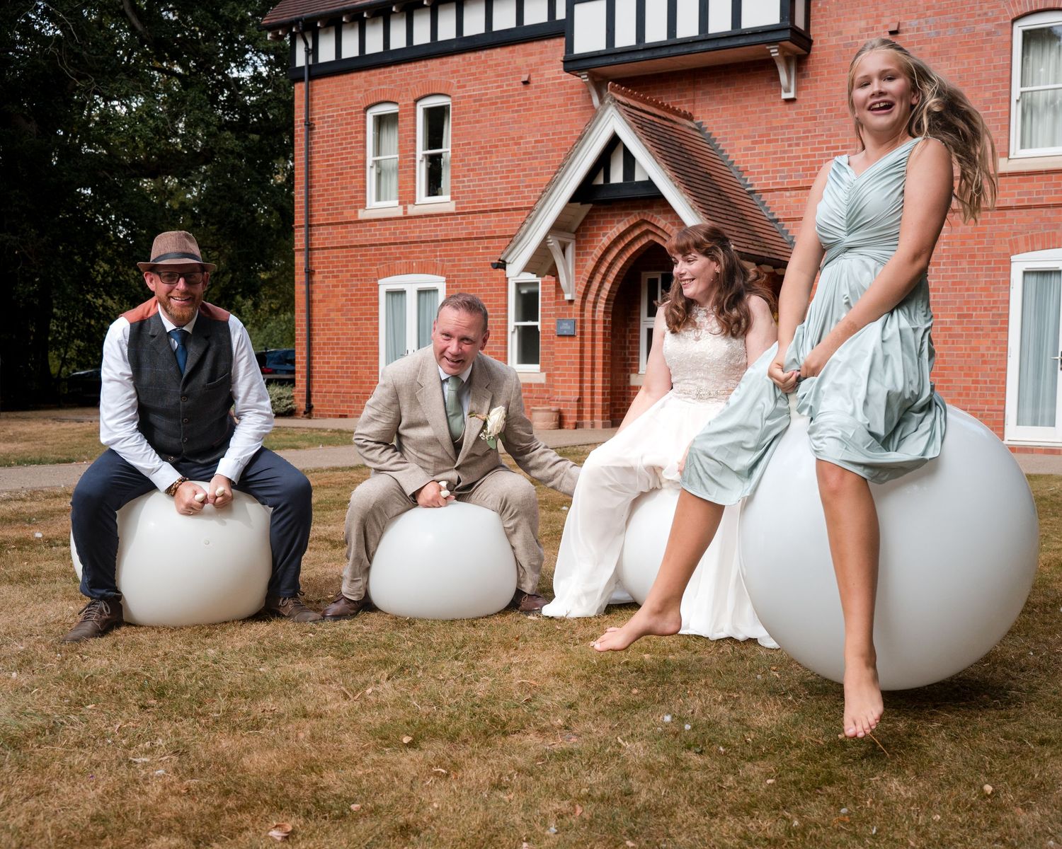 Wedding party sitting on white egg-shaped chairs outside a red brick manor house, bride in white gown with guests smiling.