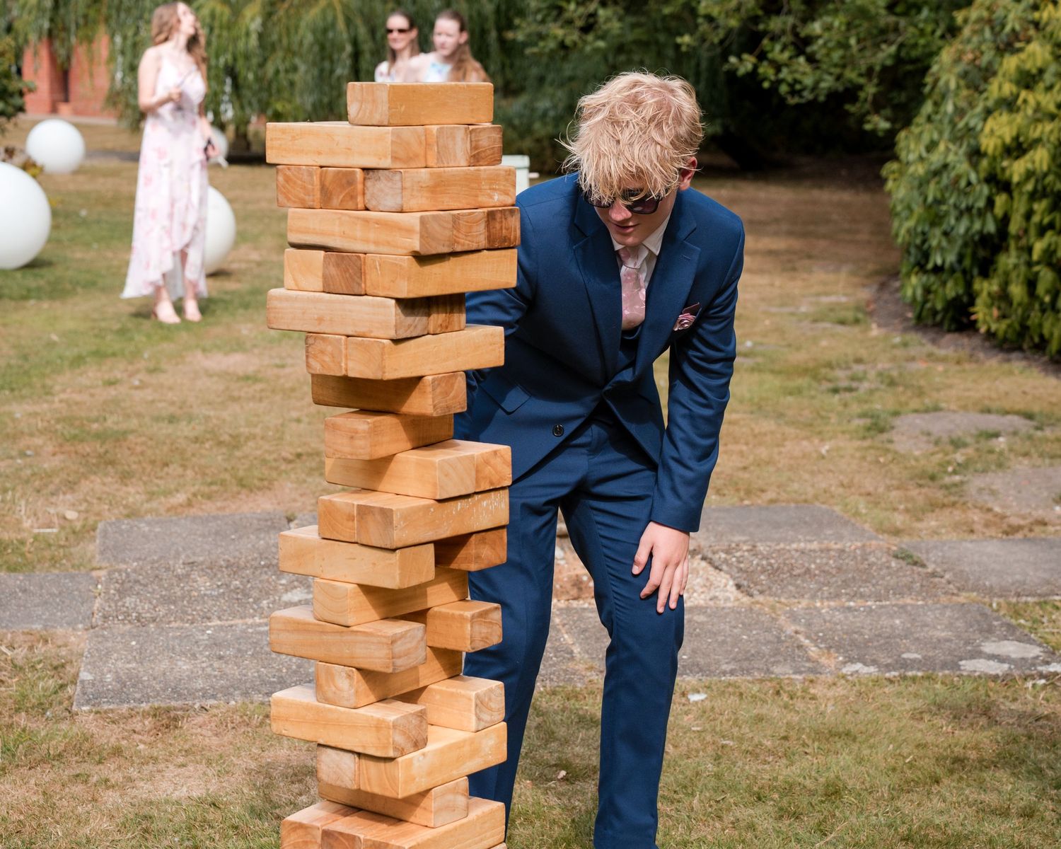 Man in blue suit playing giant Jenga outdoors at a garden party event.