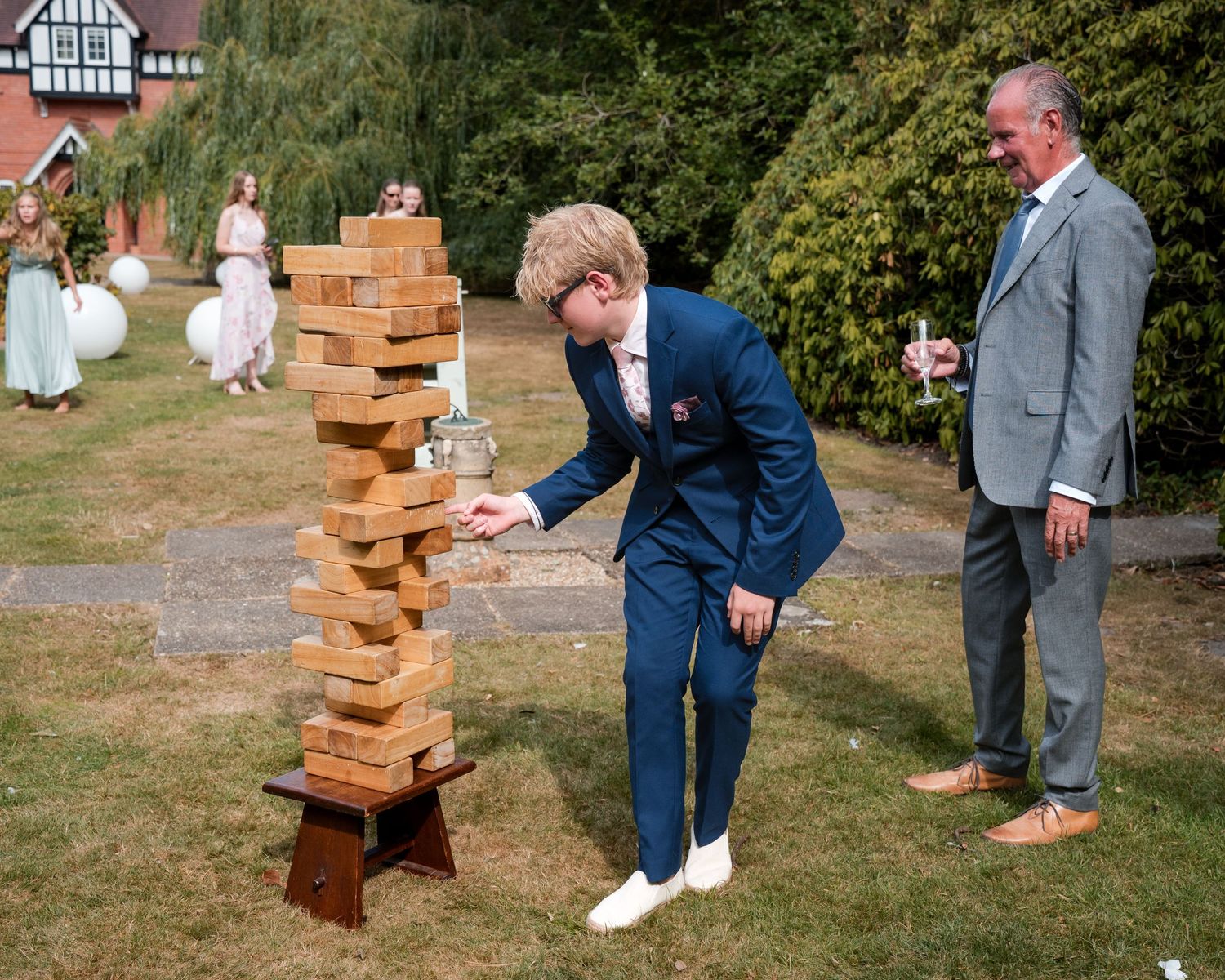 Young man in blue suit plays giant Jenga at outdoor wedding reception while older man in gray suit watches.