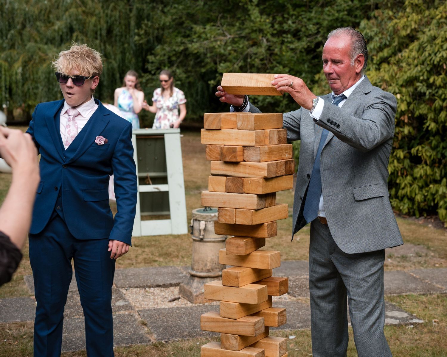 Two men in suits playing giant Jenga outdoors at a wedding reception, with guests watching in the background.