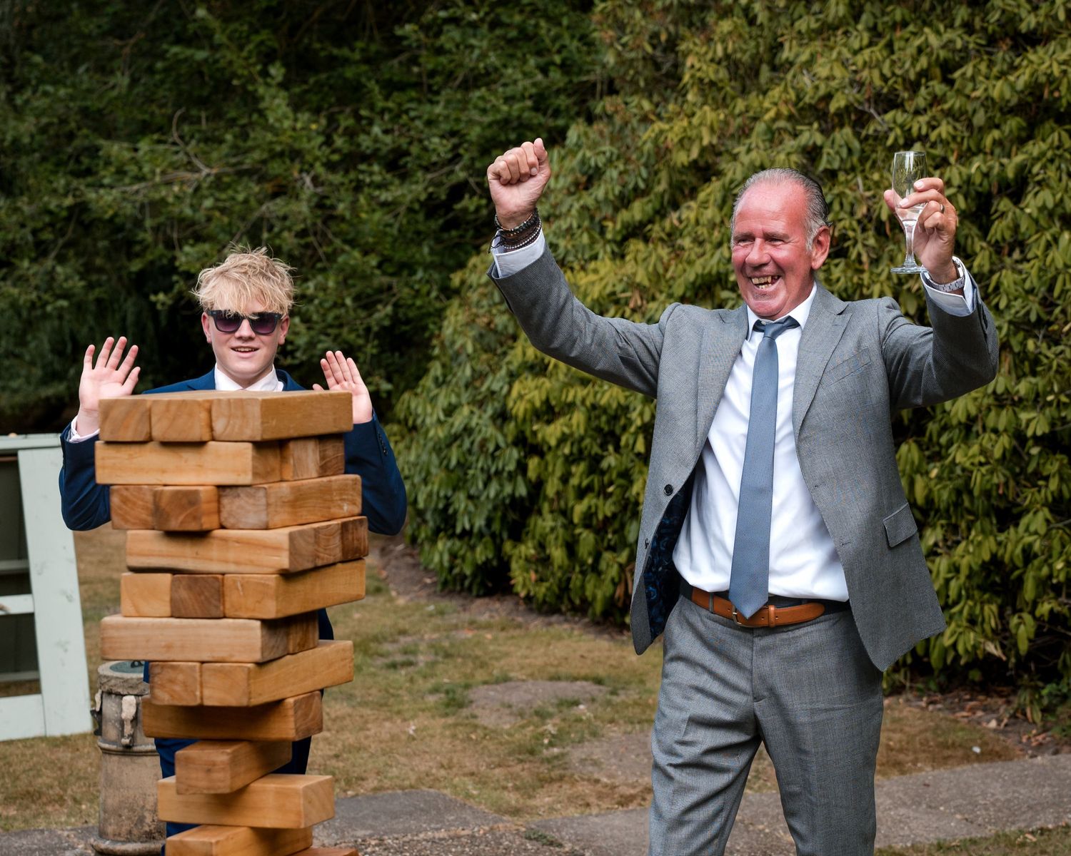 Man and boy celebrate beside giant Jenga tower at outdoor wedding reception.