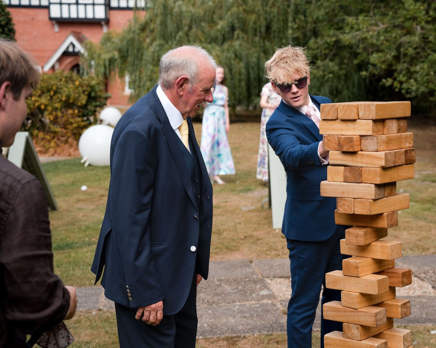 Two men in suits playing giant Jenga outdoors at a wedding reception garden party.