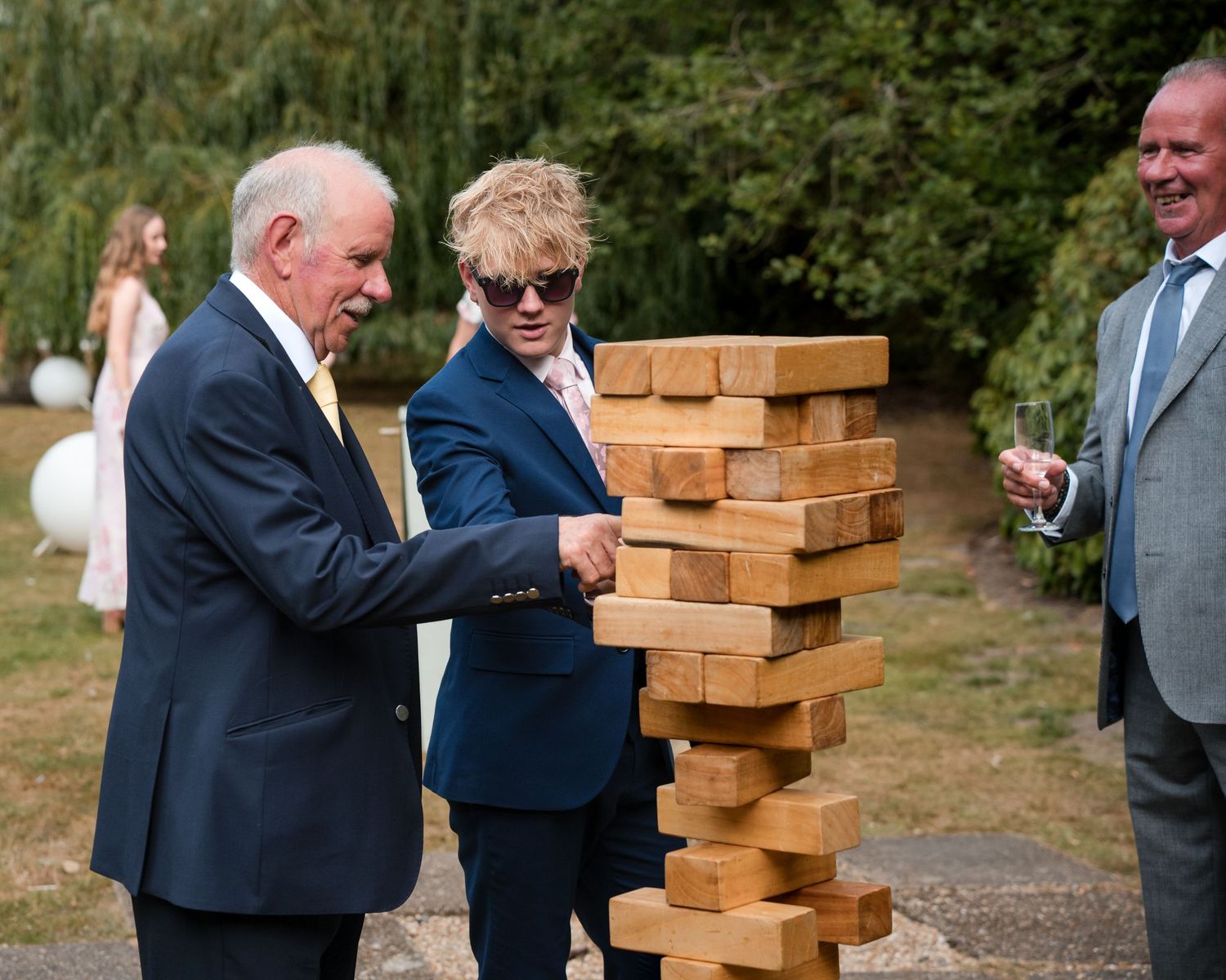 Two men playing giant Jenga outdoors at a wedding reception, with guests socializing in the background.