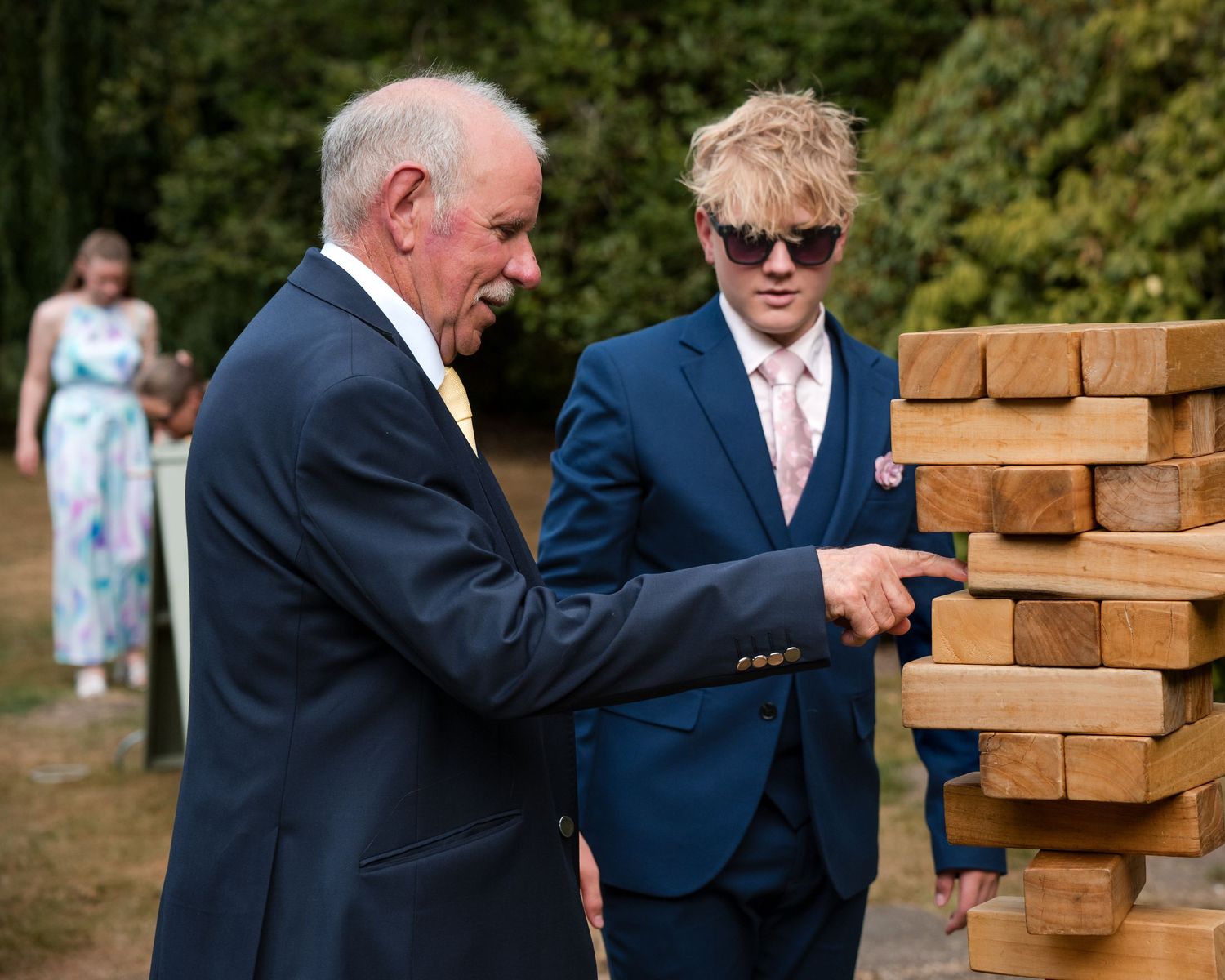 Two men in suits playing giant Jenga outdoors at a wedding reception surrounded by trees.