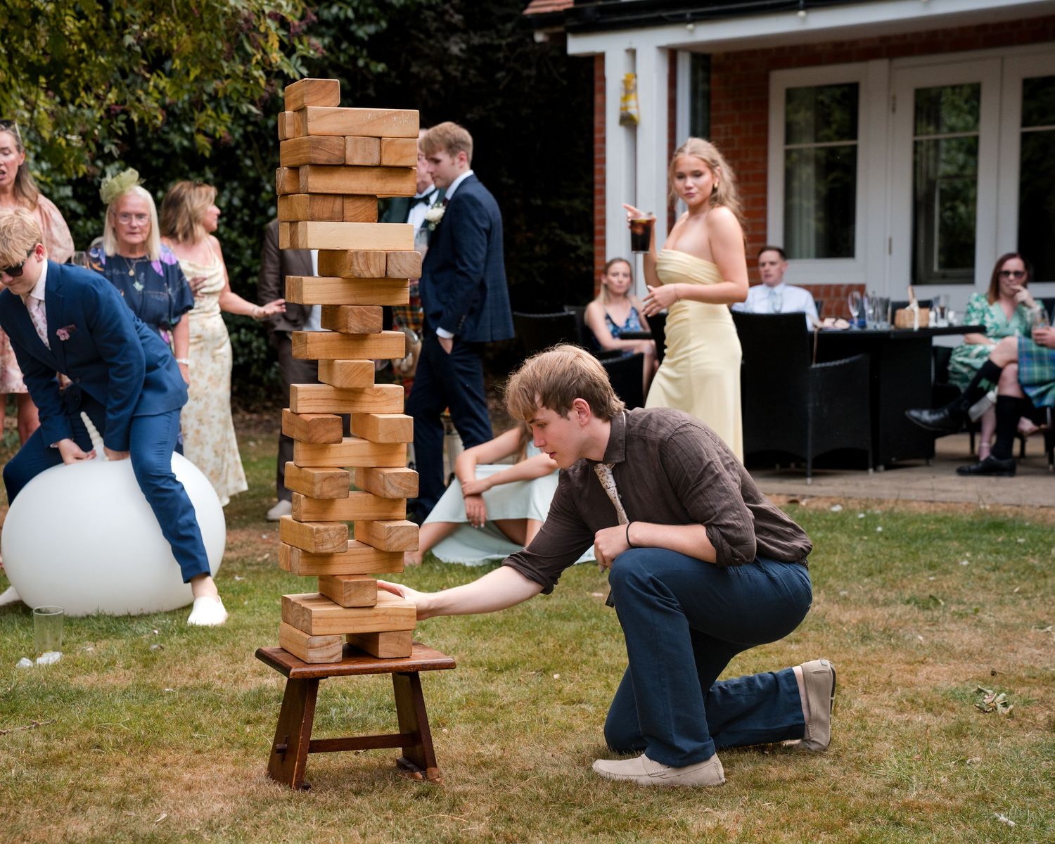 Person carefully playing giant Jenga at an outdoor wedding party with guests watching in a sunny garden.