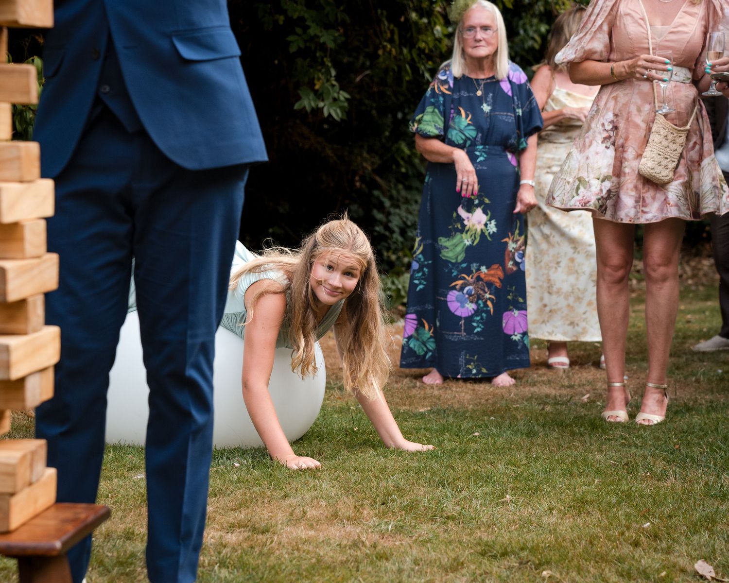 Young girl playing giant Jenga at an outdoor wedding reception while guests watch on a green lawn.