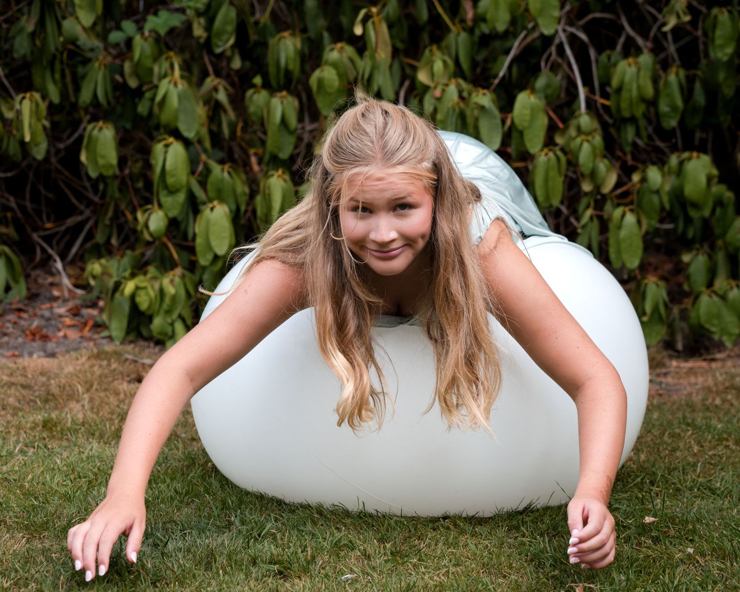Young girl with long blonde hair balancing on a white inflatable ball on green grass outdoors.