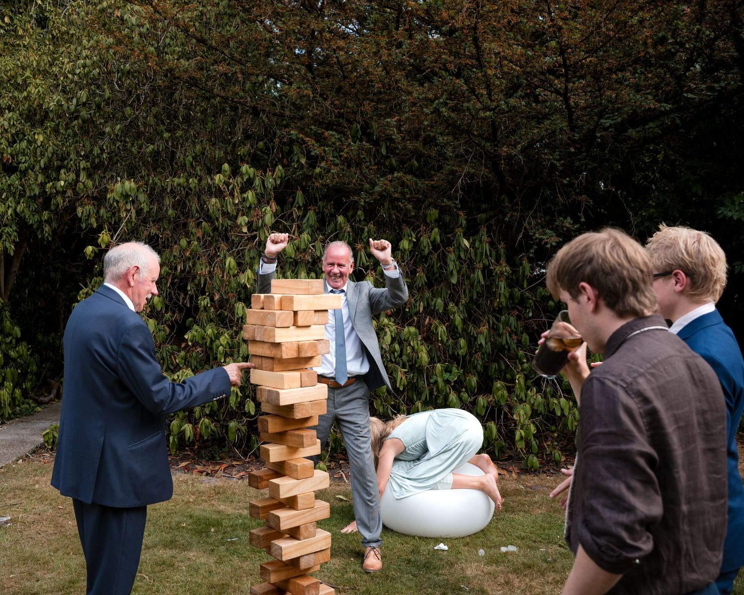 Group of people playing giant Jenga outdoors, man celebrating with raised fists as tall wooden block tower stands on grass.