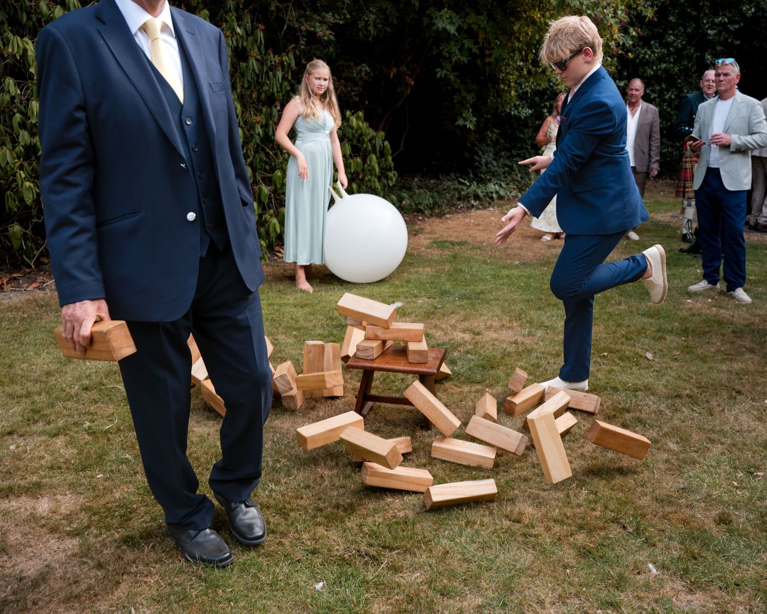People in formal attire playing giant Jenga outdoors at a wedding reception on a sunny day.