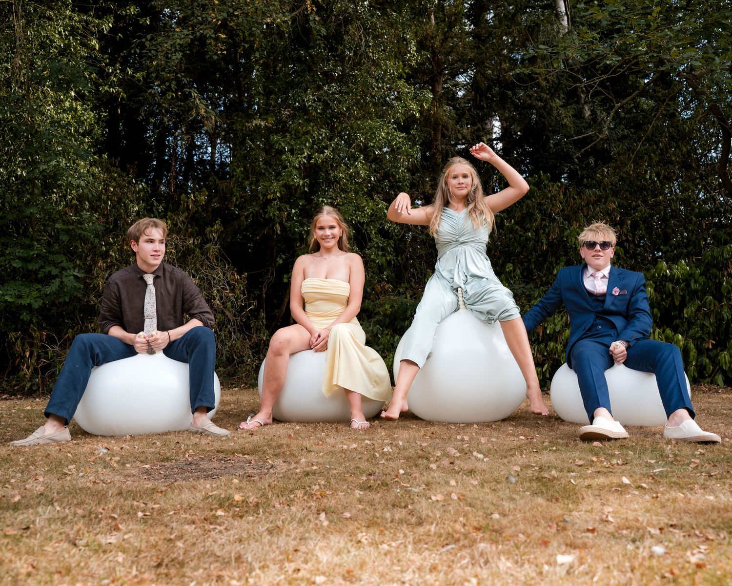 Four young adults in formal attire pose on white spherical seats outdoors with trees in the background.