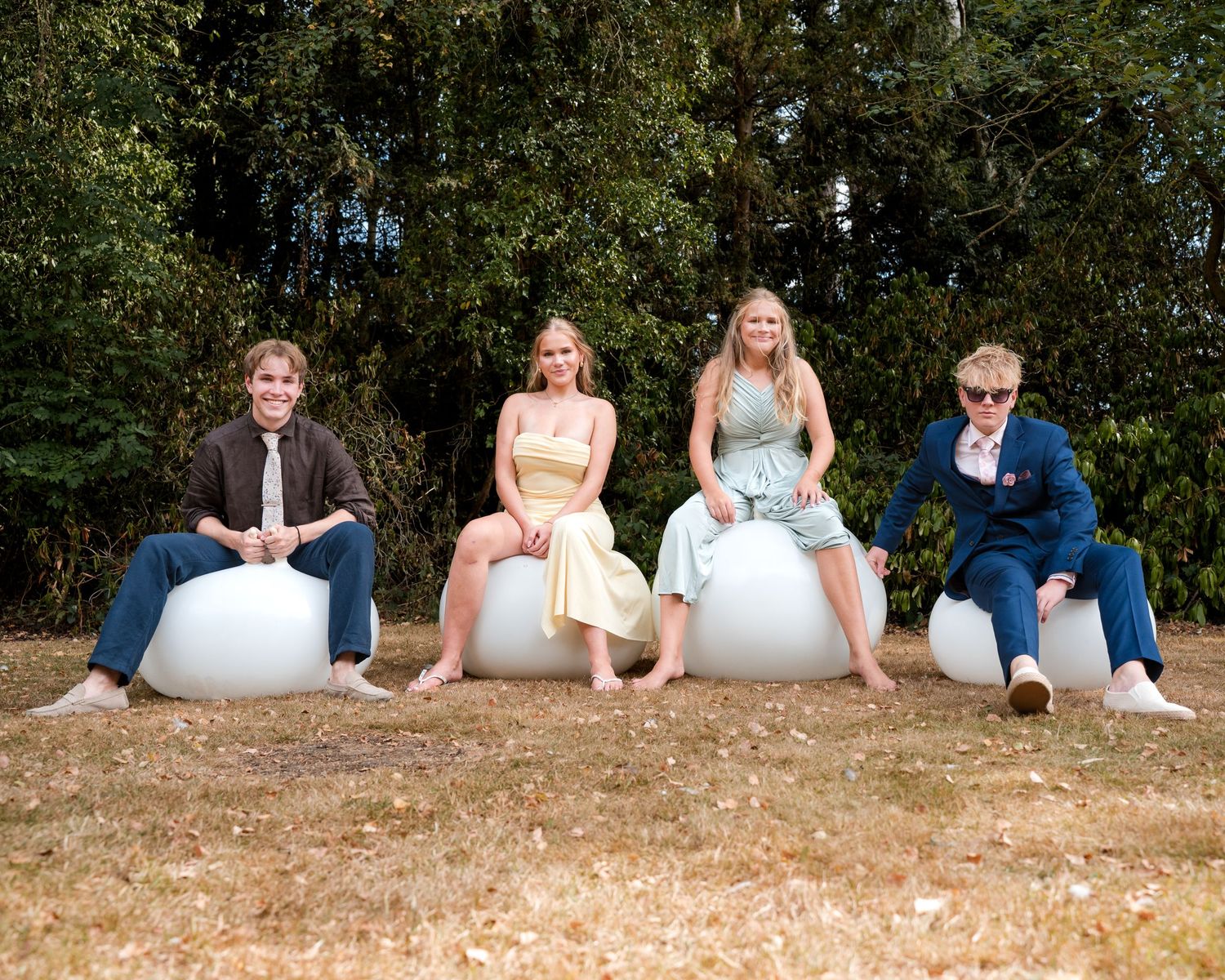 Four teens in formal attire sit on white egg-shaped seats outdoors with green trees in background.