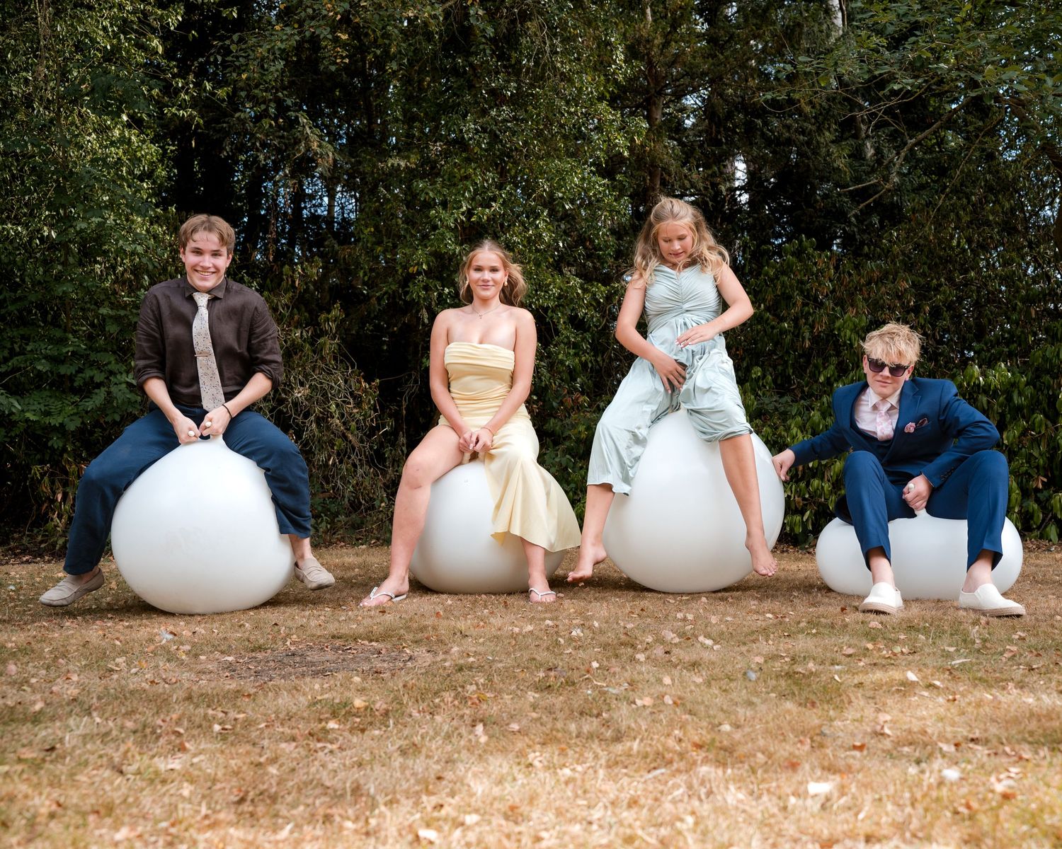 Four young people in formal attire sitting on large white spherical sculptures outdoors with trees in background.