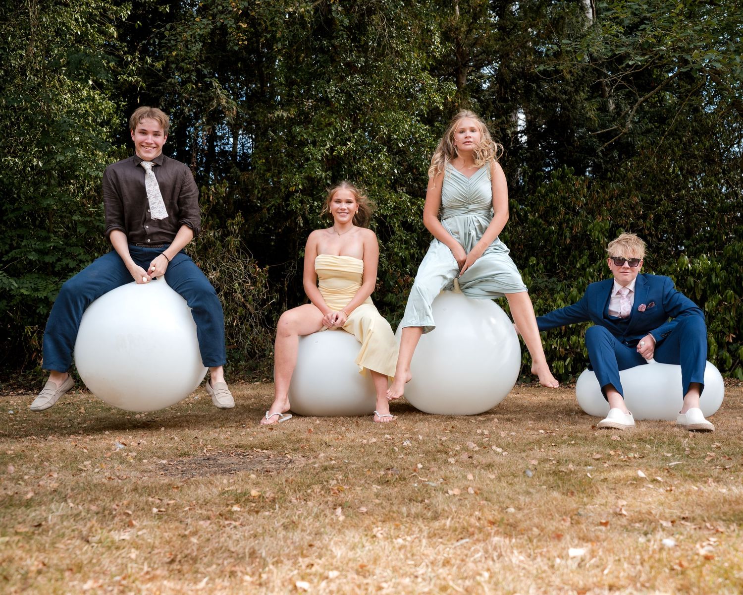 Four women sitting on large white spherical sculptures outdoors on grass with trees in background.