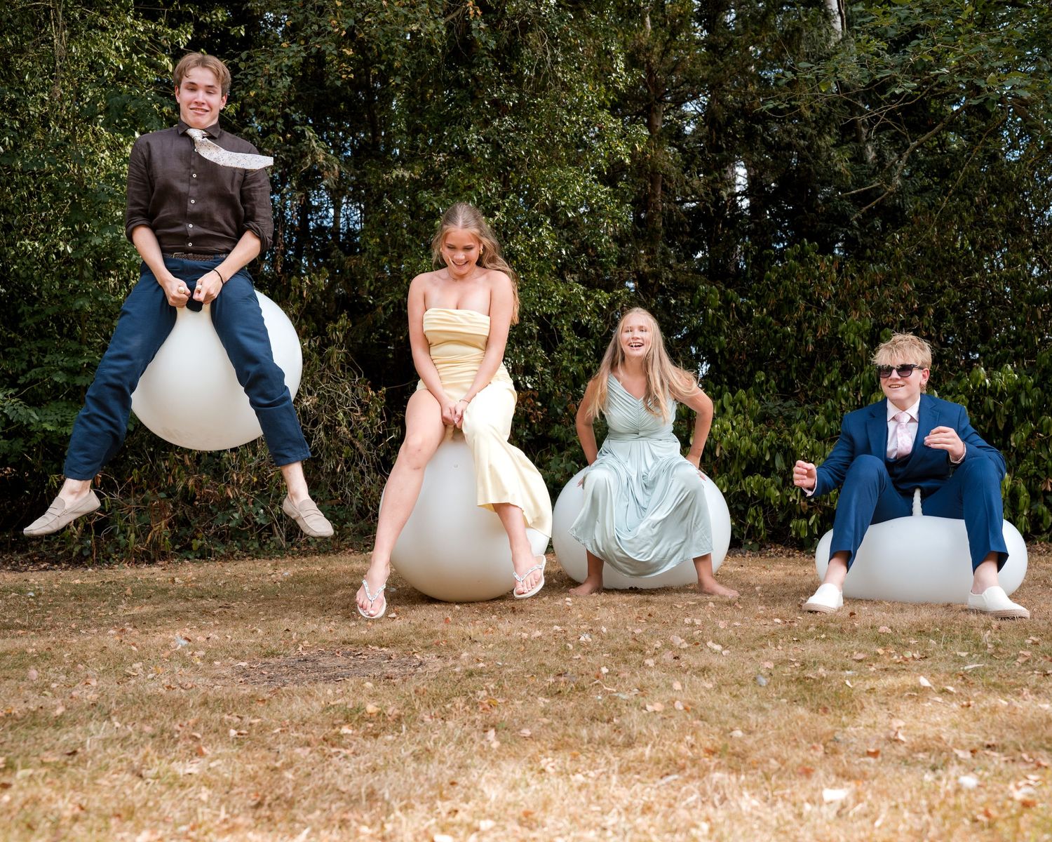Four young adults posing playfully on white space hopper balls outdoors with green trees in the background.