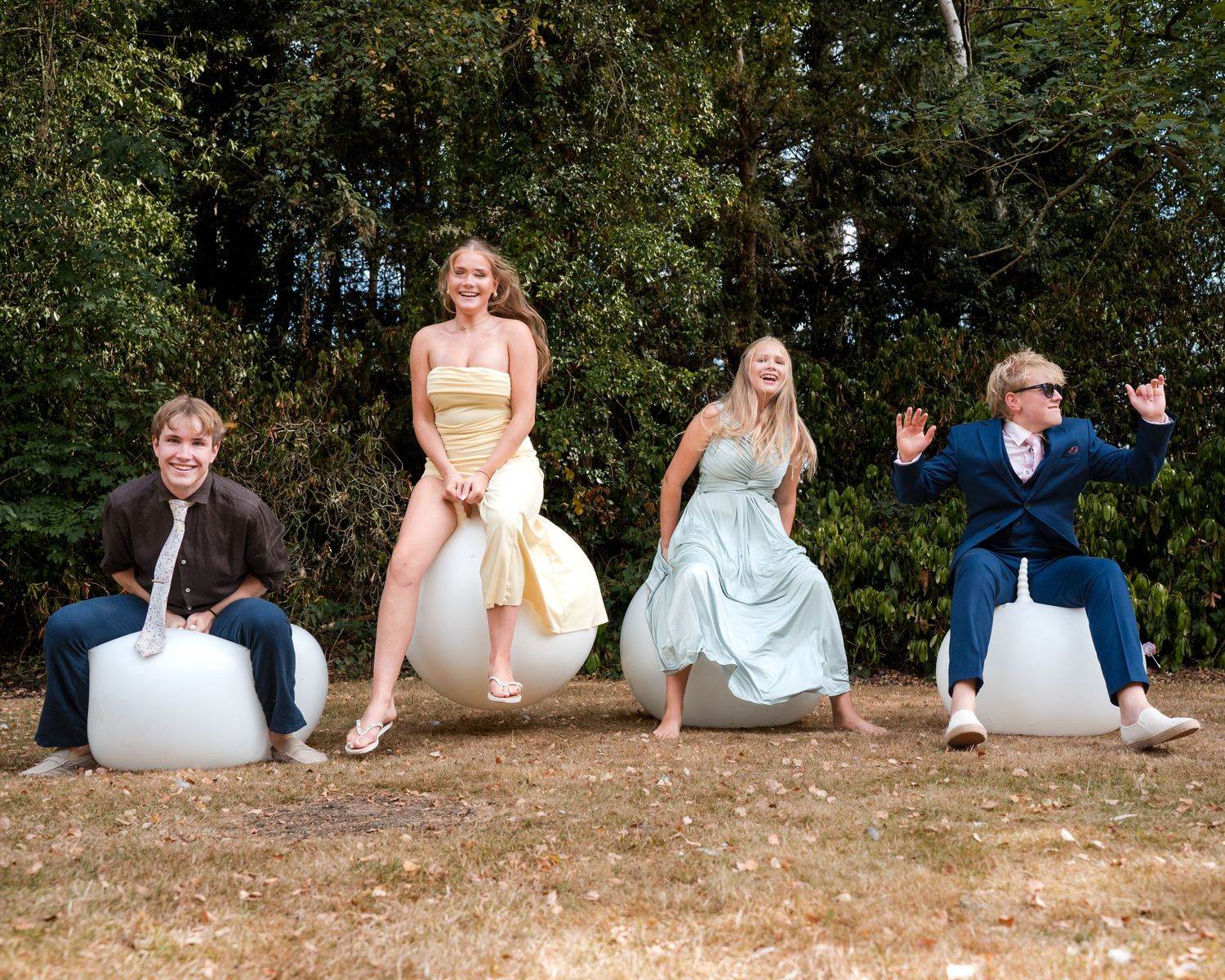 Four teens in formal attire joyfully bounce on white hopper balls outdoors with trees in background.