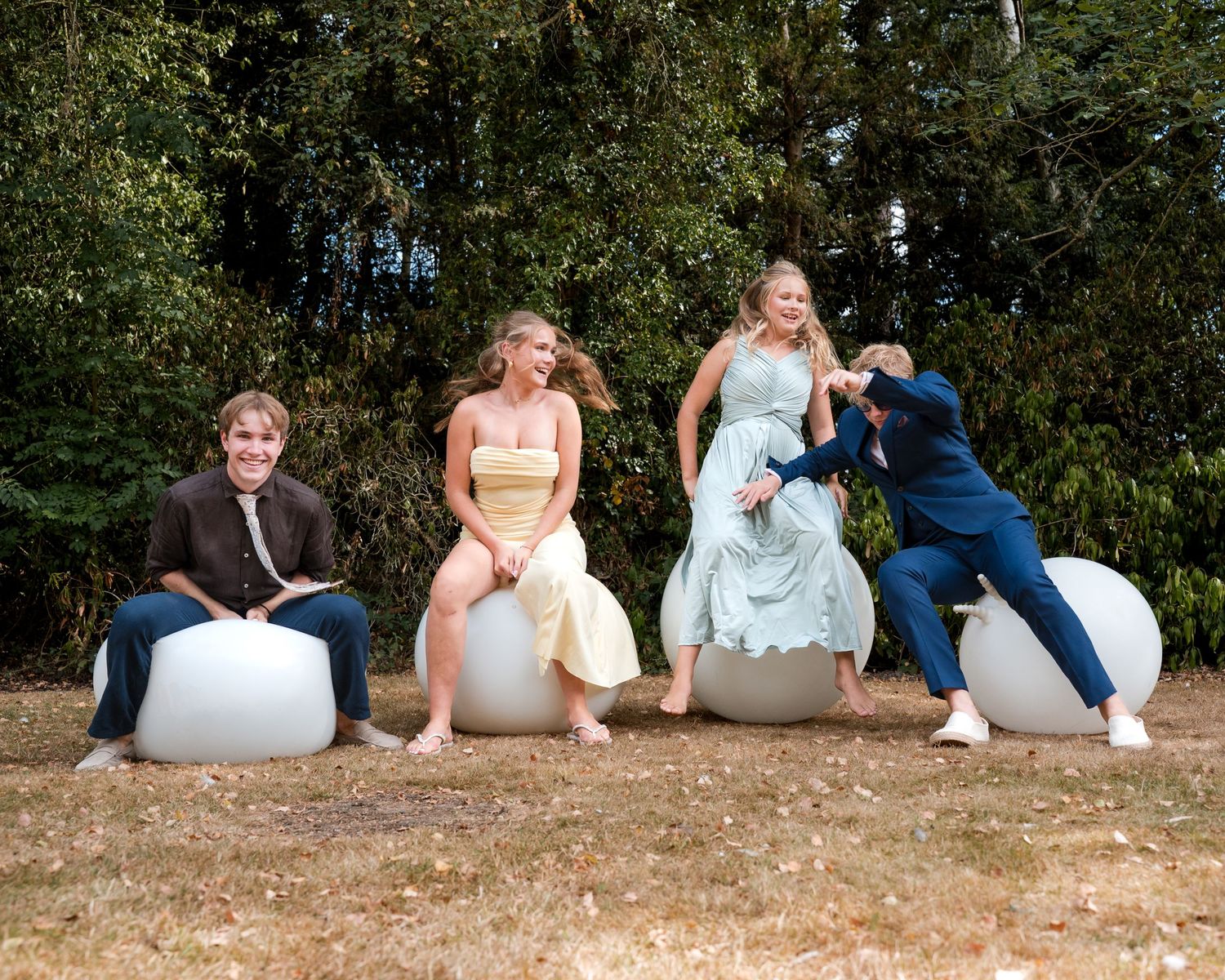 Four young adults in formal attire laugh and pose on white egg-shaped seats outdoors near lush green trees.