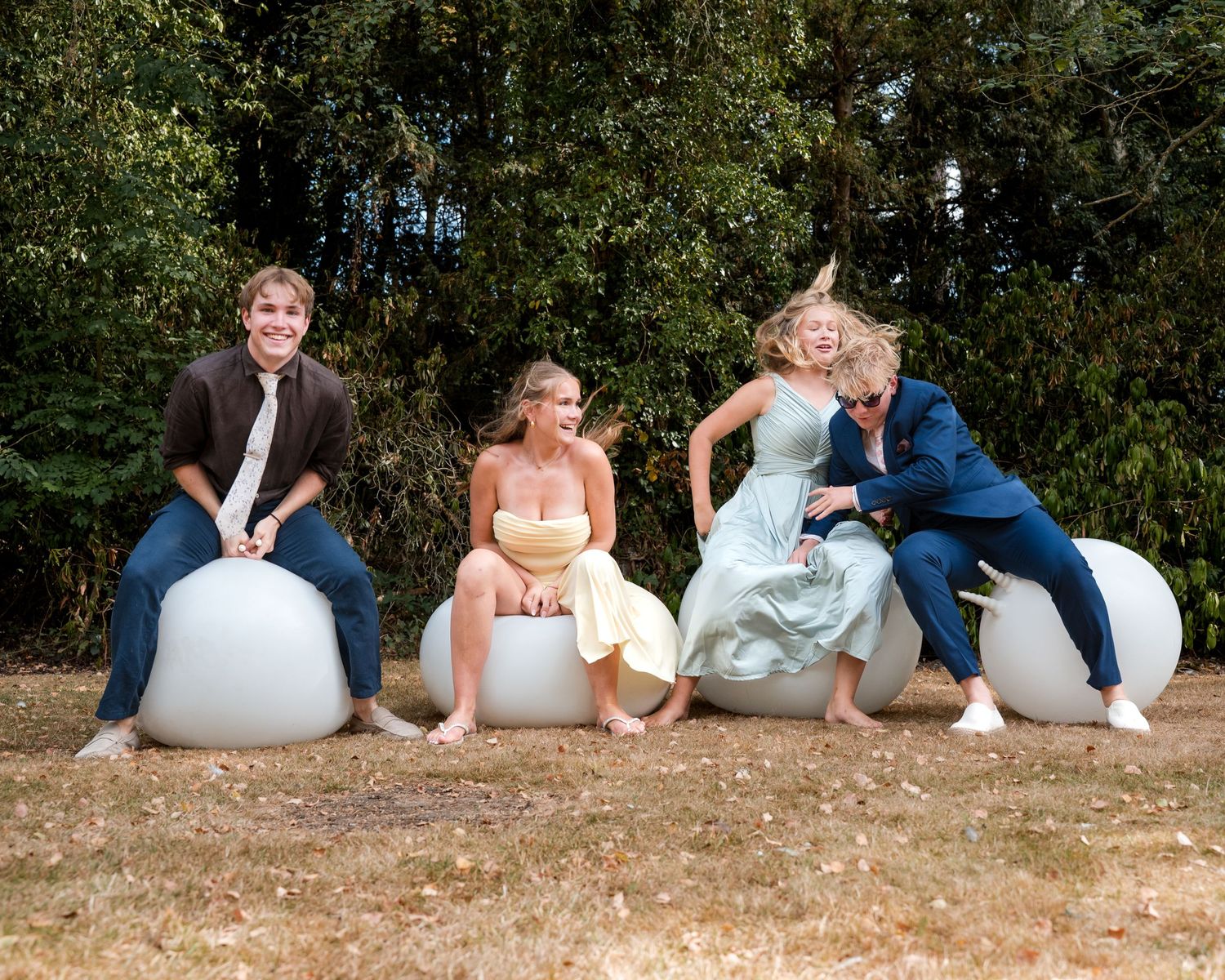 Four elegantly dressed young adults laugh and pose sitting on large white egg-shaped sculptures outdoors near trees.