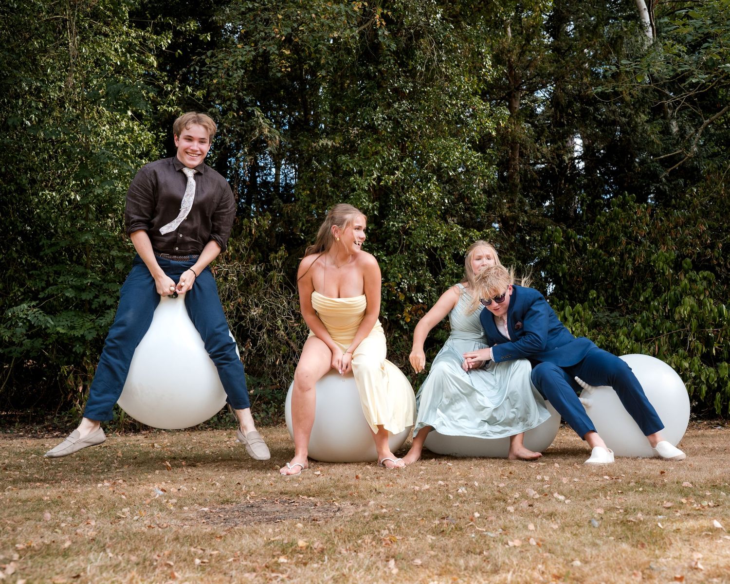 Four elegantly dressed people joyfully bouncing on large white space hoppers outdoors with trees in the background.