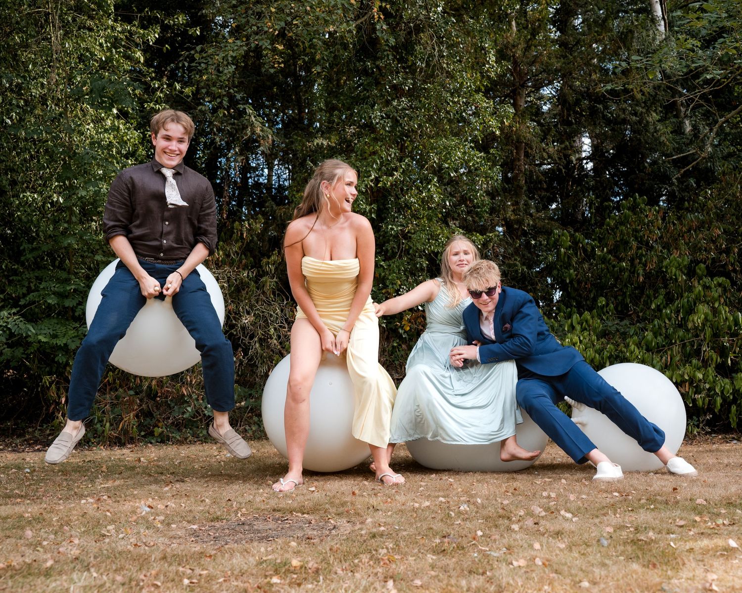 Four people in formal attire laughing and playing on large white inflatable balls outdoors.