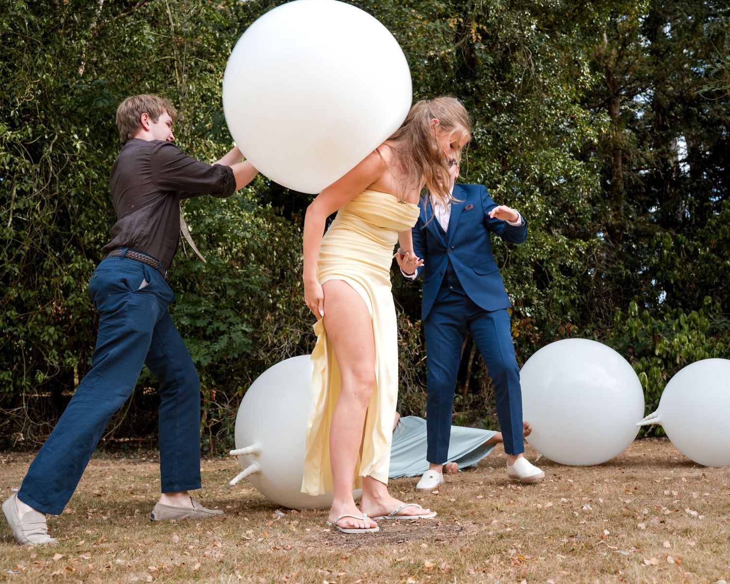 Three people playing with large white balloons outdoors at a wedding celebration on a sunny day.