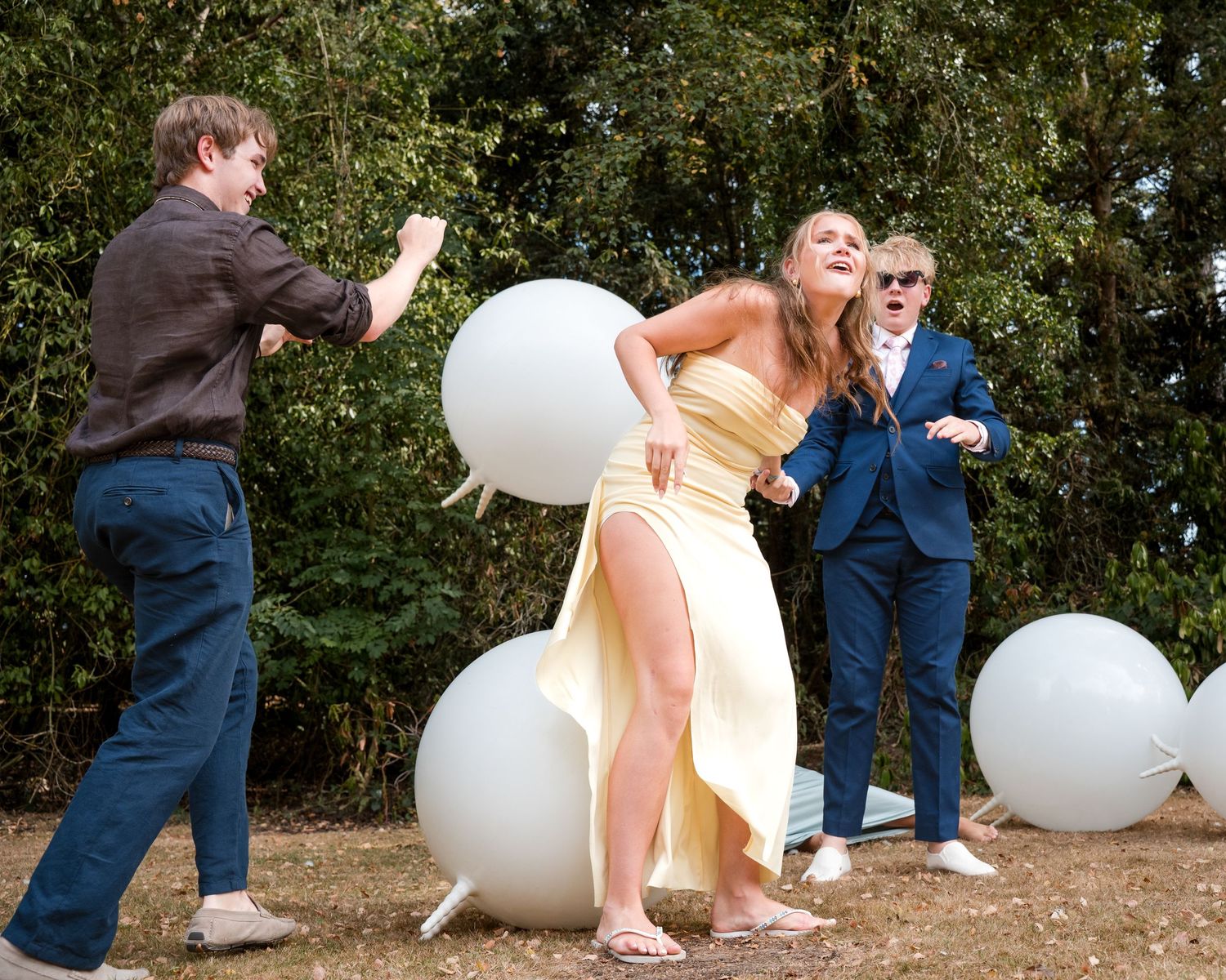 Three people dancing outdoors near large white balloons, woman in yellow dress, men in brown and blue suits.