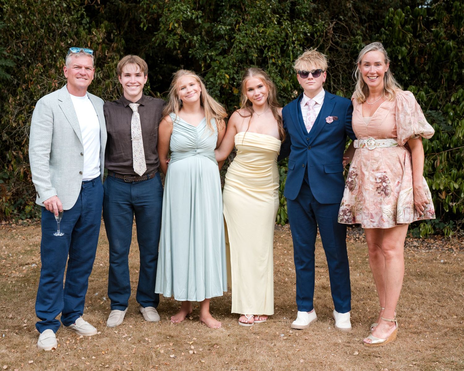 Group of six adults posing together outdoors at a formal event, dressed in elegant summer attire.