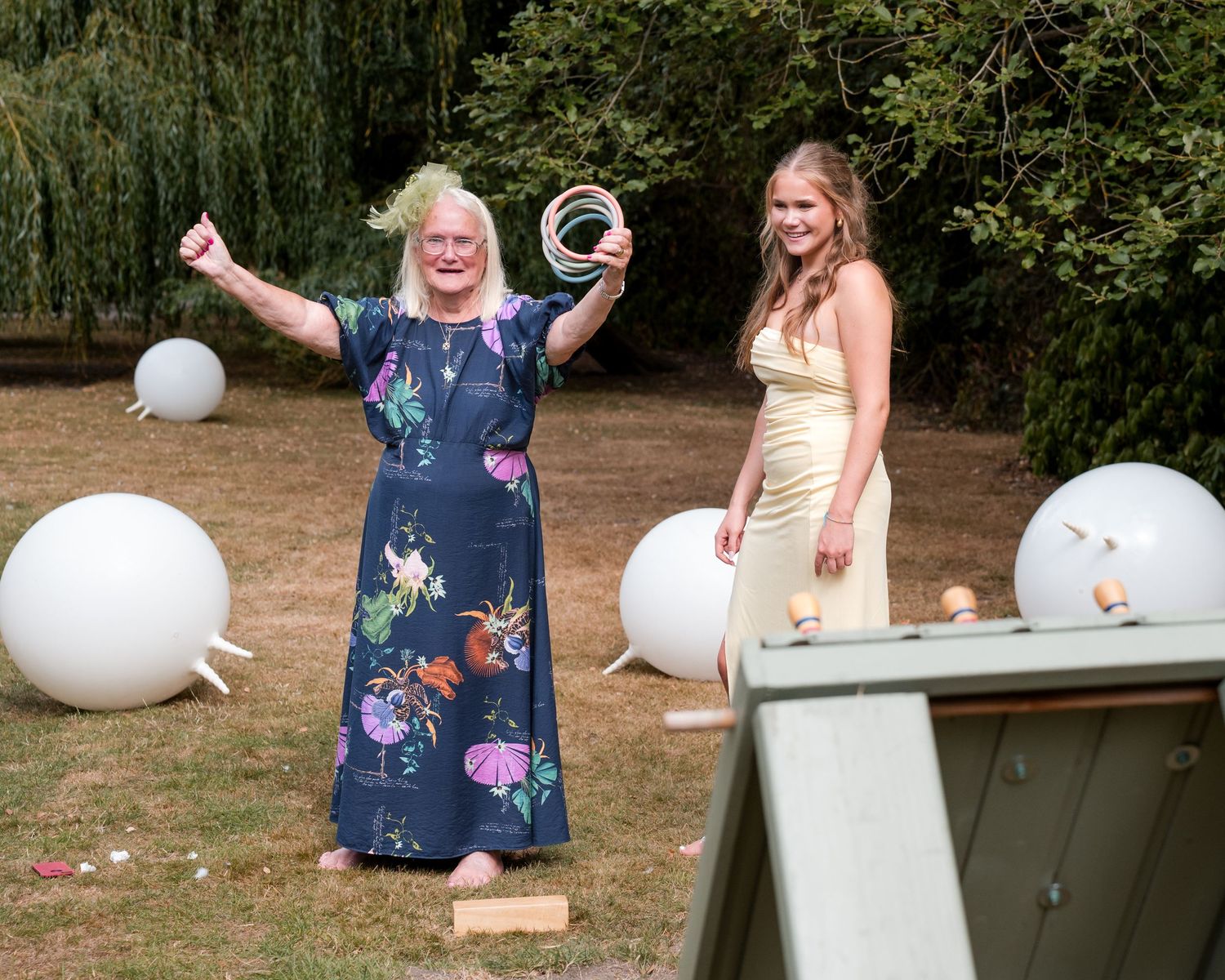 Two women playing ring toss lawn game outdoors at a garden party, surrounded by white decorative balls.