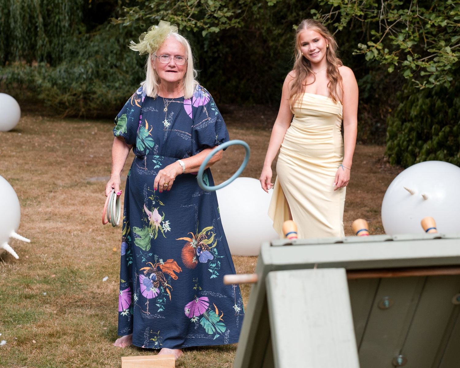 Two women in elegant dresses pose outdoors at a garden party with white balloon decorations and lush greenery.