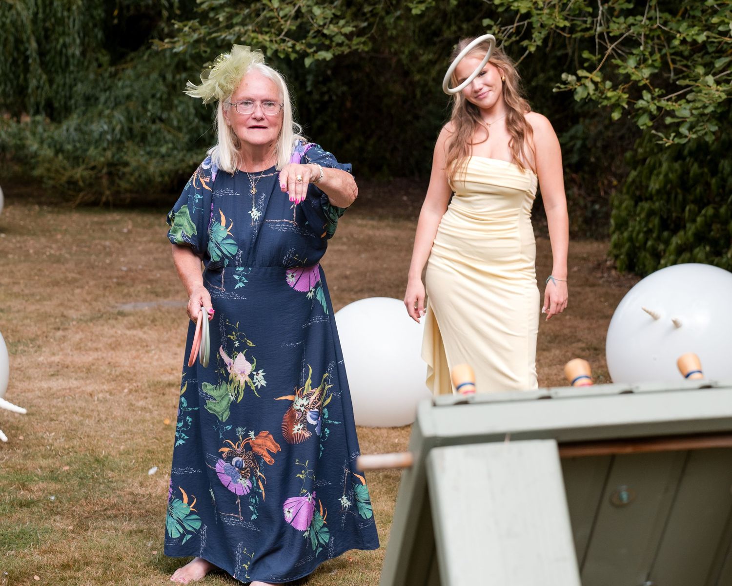 Two women in elegant dresses stand outdoors at a garden party, one in floral blue and one in yellow gown.
