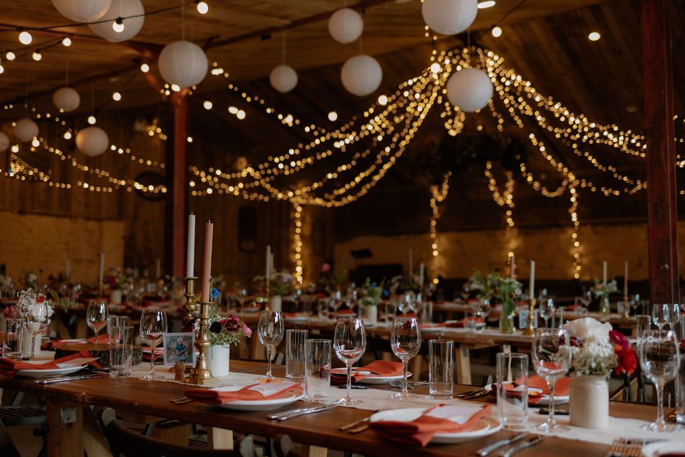 Rustic barn wedding reception with string lights, white lanterns, long wooden tables, and elegant place settings with red napkins.