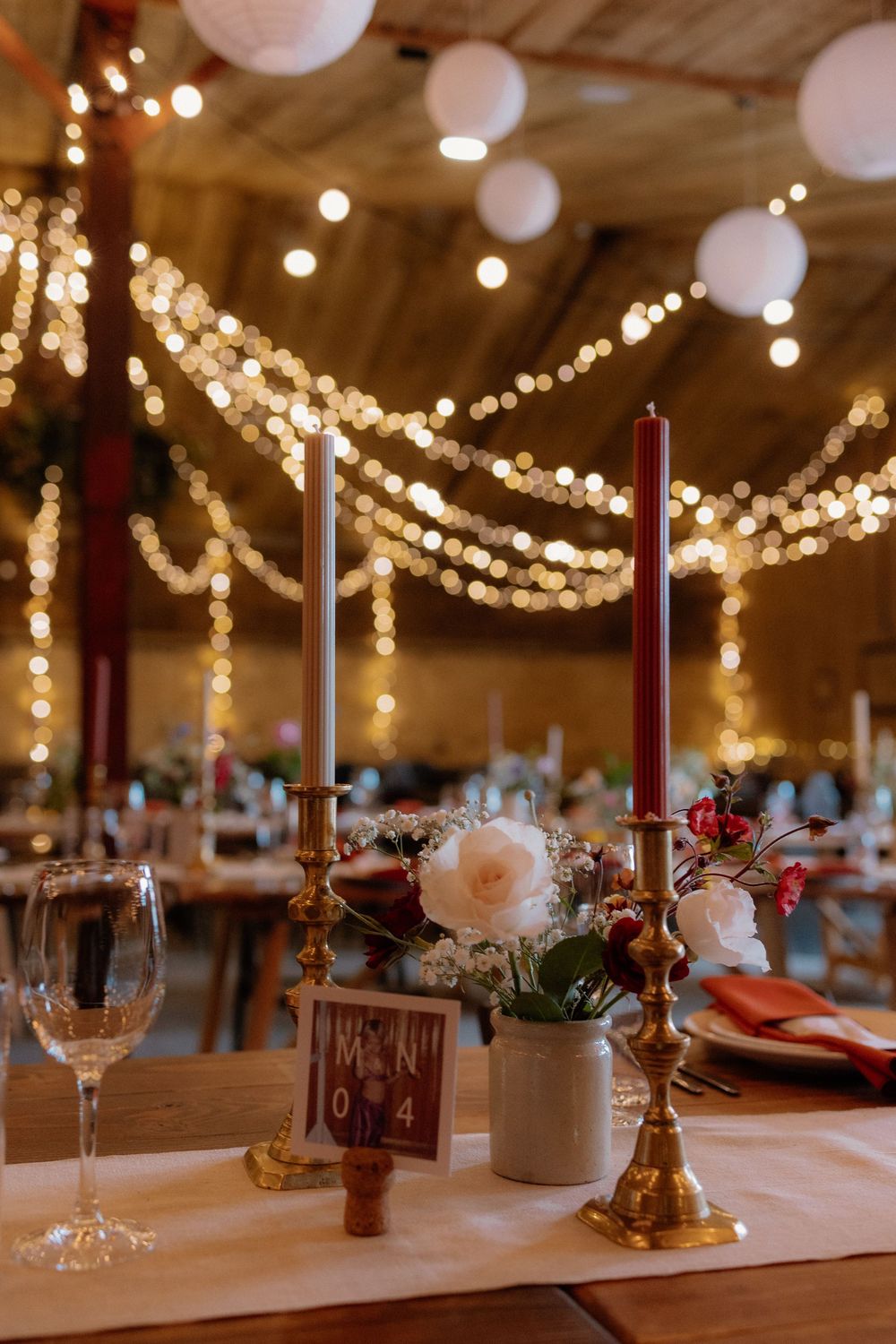 Elegant wedding reception table with gold candlesticks, white candles, floral centerpiece, and string lights in a warm barn venue.