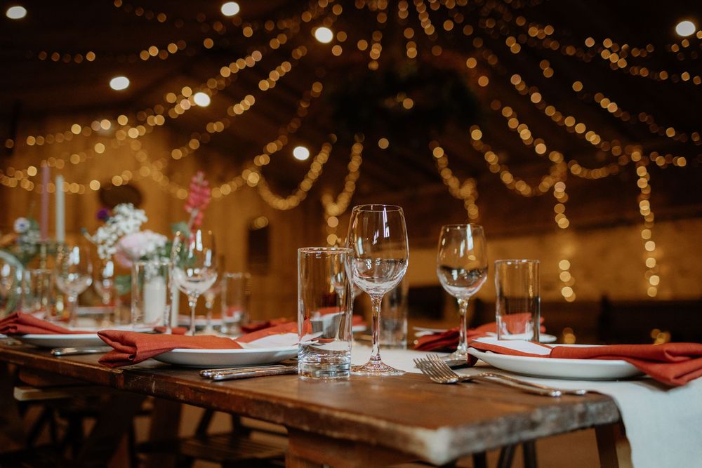 Elegant wedding reception table set with crystal glasses, red napkins, and warm string lights in a rustic barn venue.