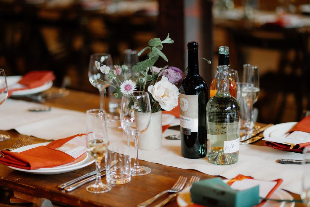 Elegant wedding reception table with orange napkins, wine bottles, floral centerpiece, and glassware on a wooden table.