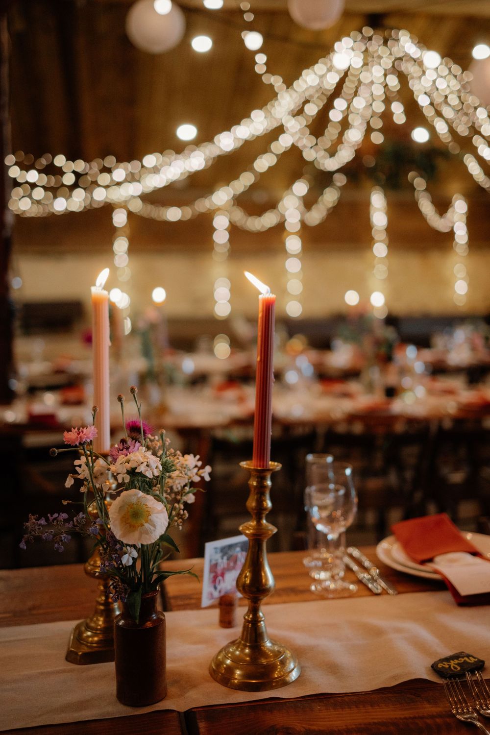 Elegant wedding reception with brass candlesticks, wildflower arrangements, and twinkling string lights in a warm barn venue.