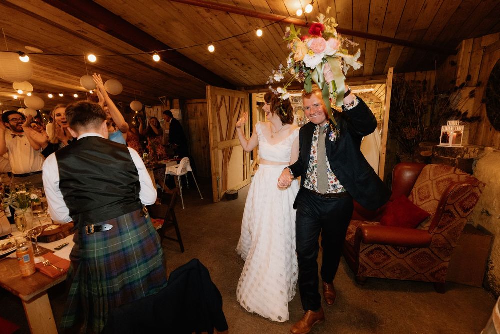 Bride and groom dancing joyfully at rustic wedding reception, groom wearing tartan kilt, bride holding floral bouquet overhead.