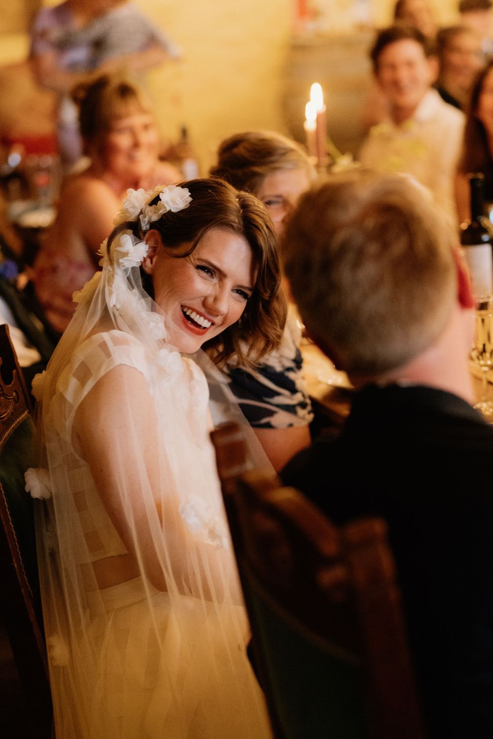 Smiling bride in floral veil laughing with guest at wedding reception dinner table with candlelight.