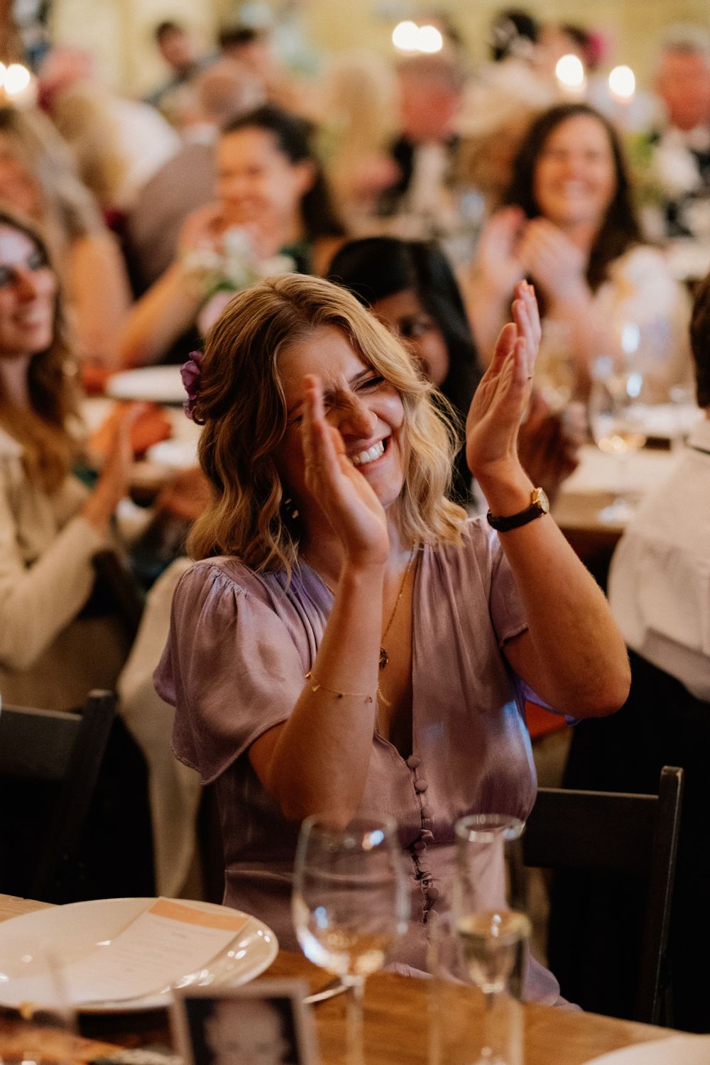 A joyful wedding guest in lavender dress laughing and clapping at a reception dinner celebration.