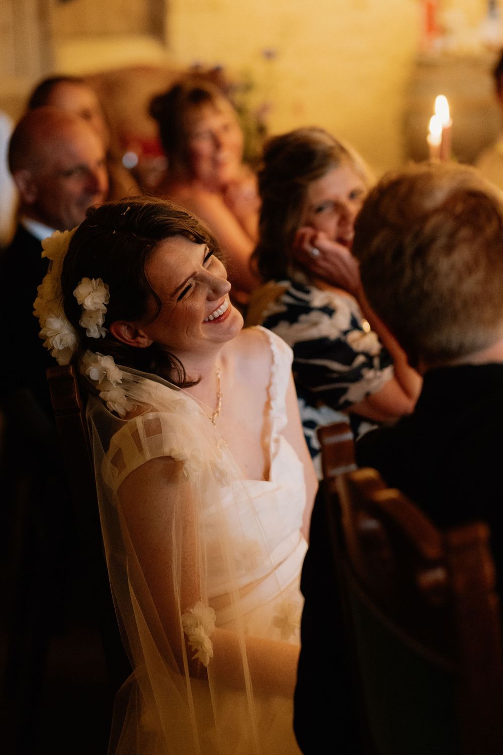 Radiant bride smiling joyfully during wedding ceremony, wearing white dress and floral hair accessories, surrounded by guests.