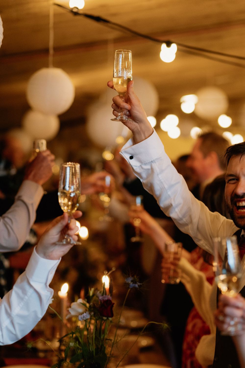 Guests raising champagne glasses in a toast at an elegant candlelit celebration with string lights and white balloon decor.