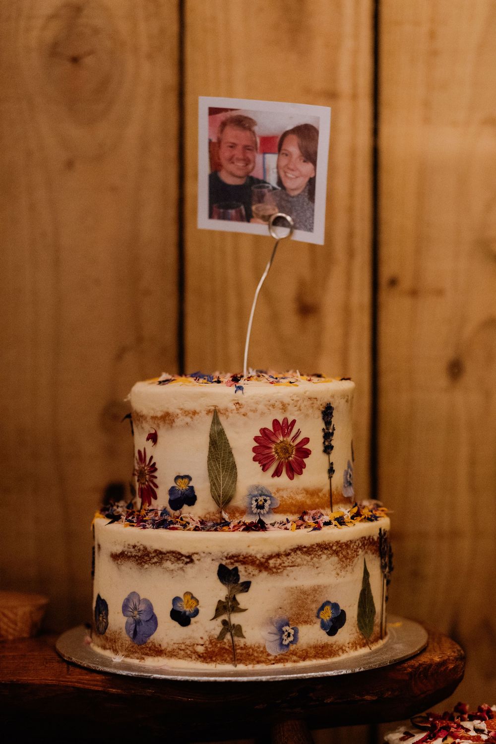 Two-tier white wedding cake decorated with pressed flowers and a polaroid photo topper on a wooden background.