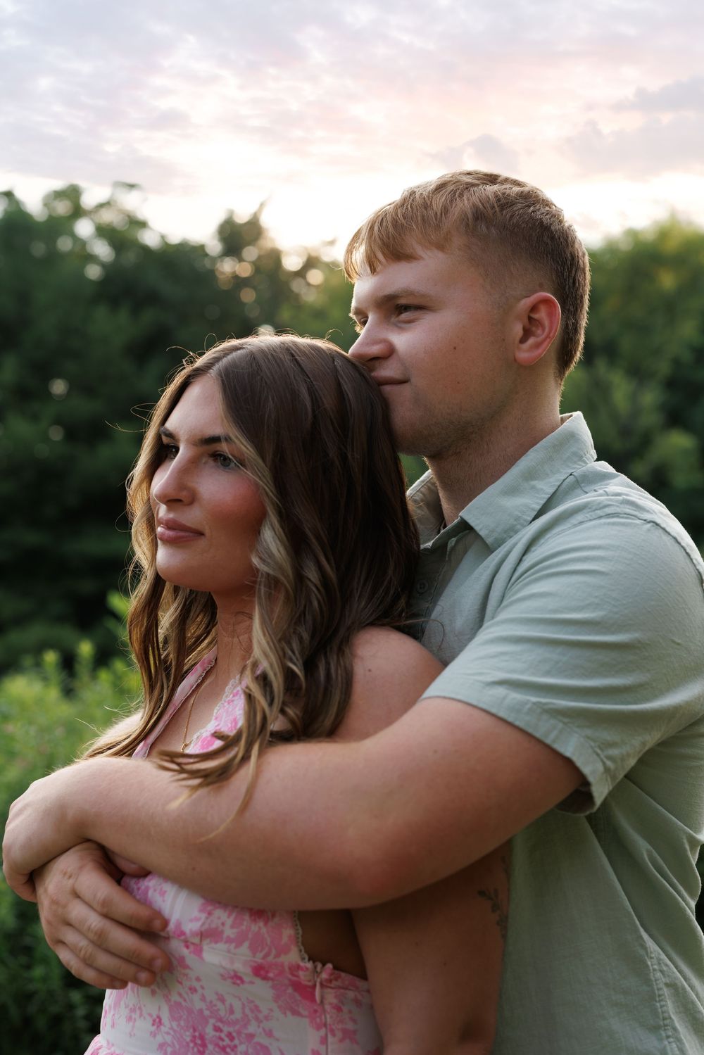 A couple embraces outdoors at sunset, man in green shirt hugging woman in pink floral dress with trees behind them.
