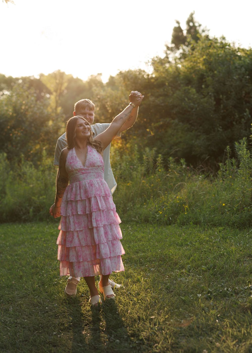 Couple dancing outdoors at golden hour, woman in pink ruffled dress, man behind her raising their joined hands.