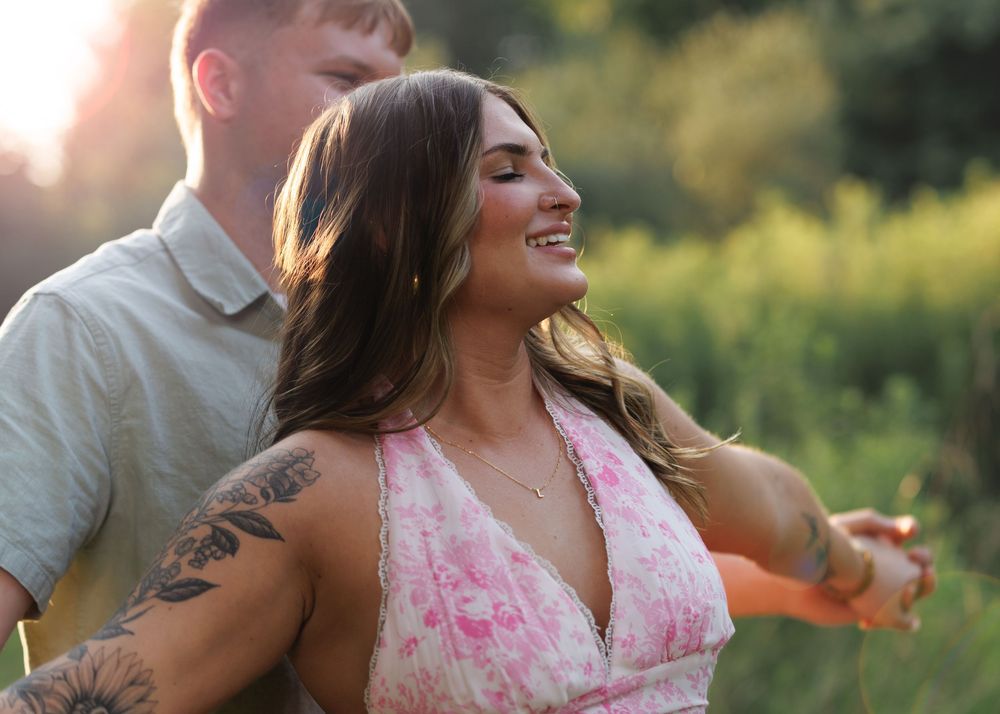Smiling woman in pink floral dress with tattoos enjoys a sunny outdoor moment with a man in a green field.