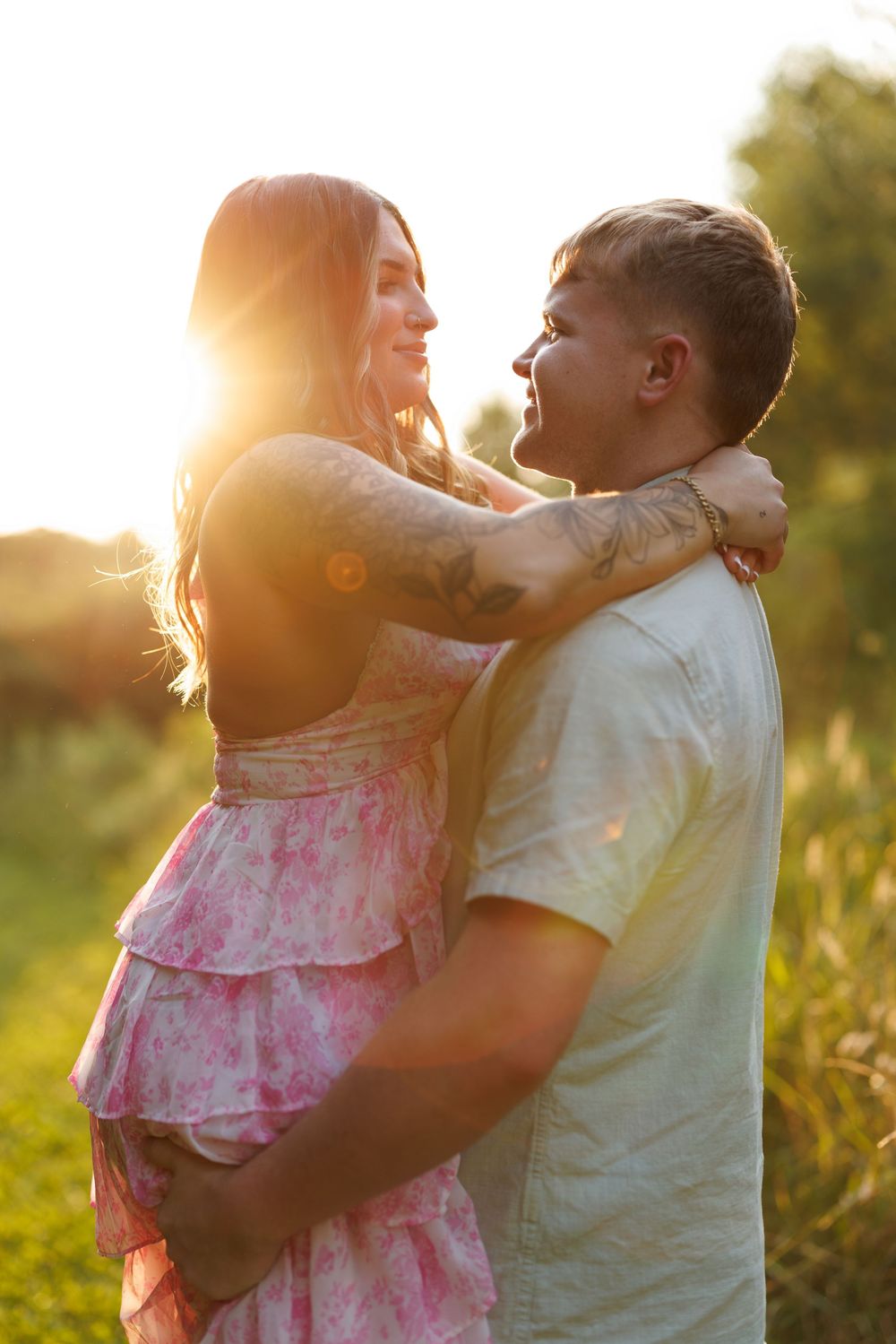 Couple embracing at golden hour sunset in a field, woman in pink floral dress with tattoos, romantic outdoor portrait.