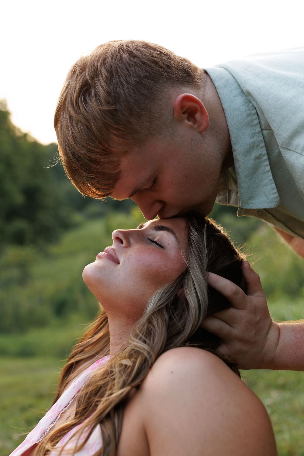 A young couple shares a tender kiss outdoors, with the man gently cradling the woman's head in a green field.