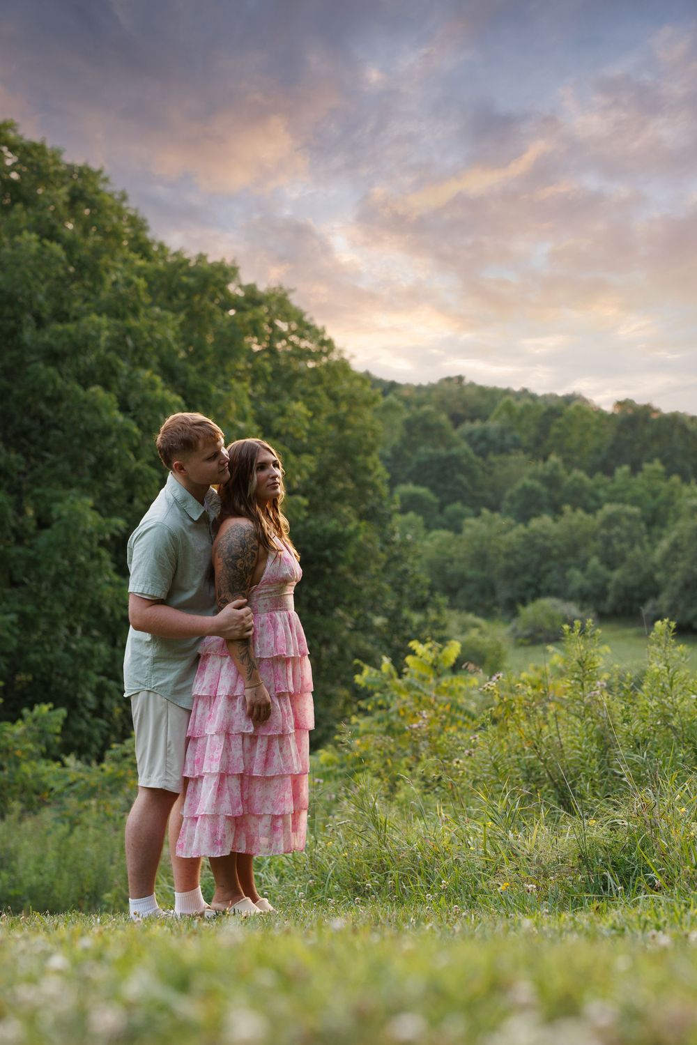 Couple embracing in a lush green meadow at sunset, woman in pink ruffle dress, man in gray shirt.
