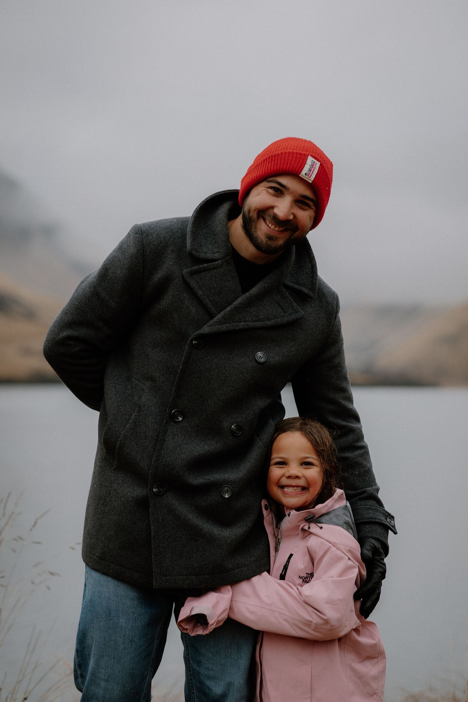 A smiling man in a red beanie and gray coat poses with a young girl in a pink jacket near a misty lake.