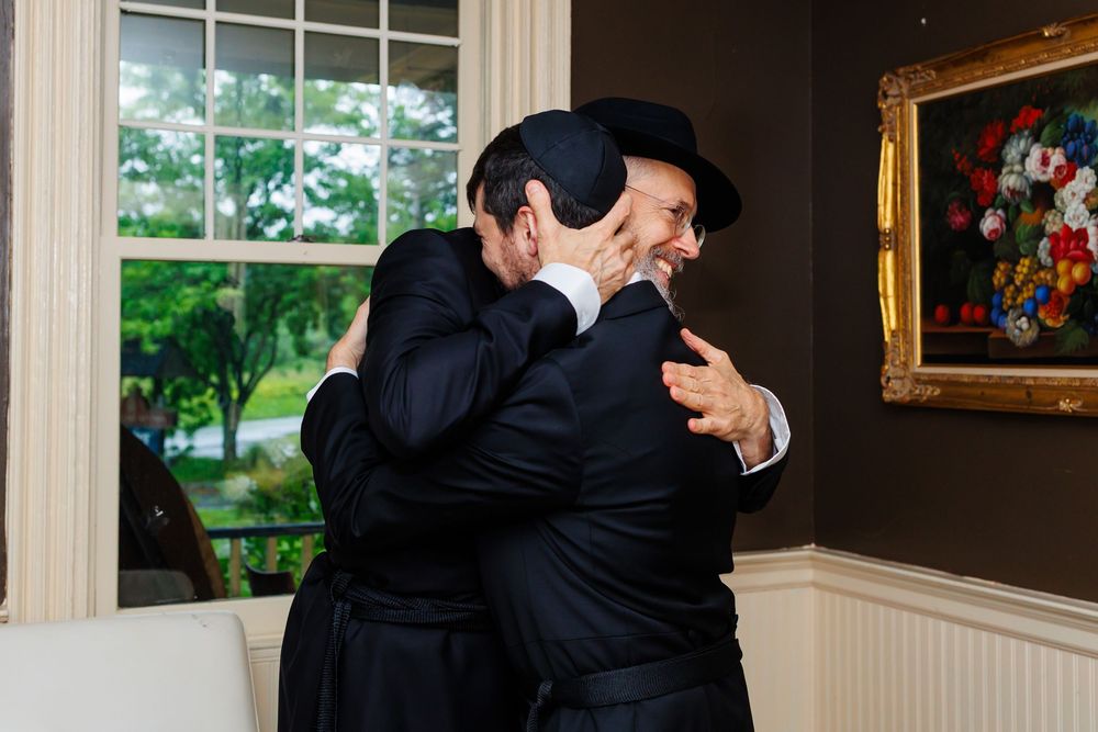 Two men in black suits share a warm embrace near a window with garden views and floral artwork on the wall.